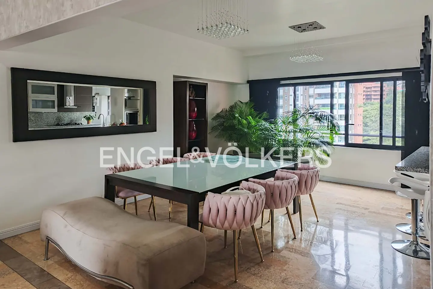 A dining room with a glass table, pink chairs, a bench, and a large mirror reflecting the kitchen.