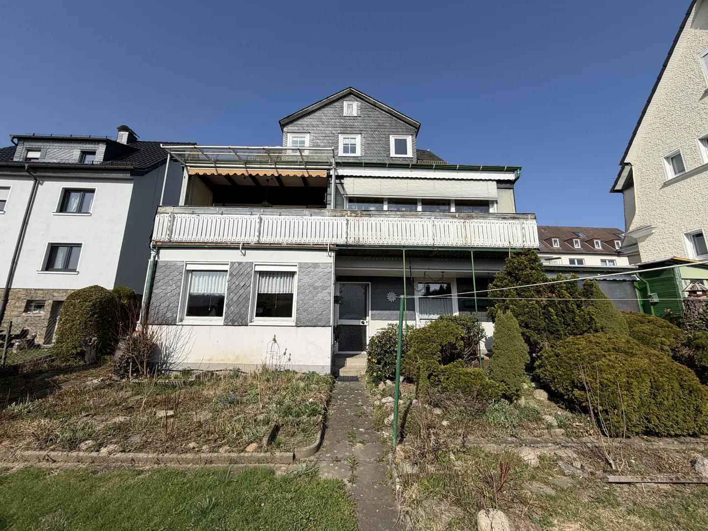 Two-story house with balconies, gray slate siding, and a stone pathway leading to the front door. Green bushes and trees surround the house.