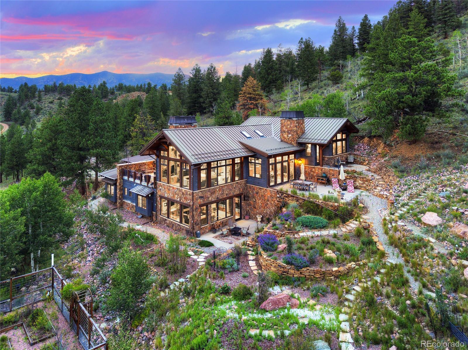 Modern mountain home with stone exterior and metal roof, nestled in a lush forest at sunset with terraced landscaping. [AI-generated text]