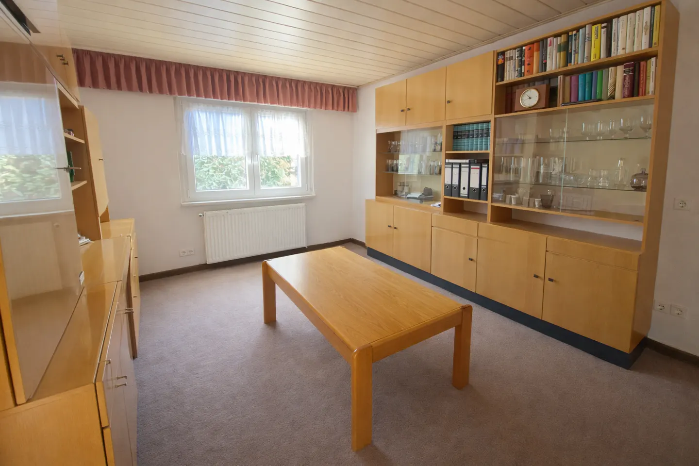 A living room with a light brown wooden table, a large wooden cabinet with books and glass objects, and a window with white curtains.