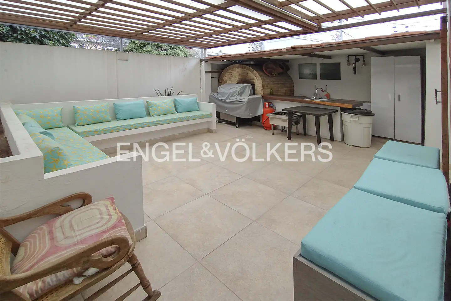 Outdoor patio with built-in seating, turquoise cushions, wood pergola, brick oven, and Engel & Völkers logo.