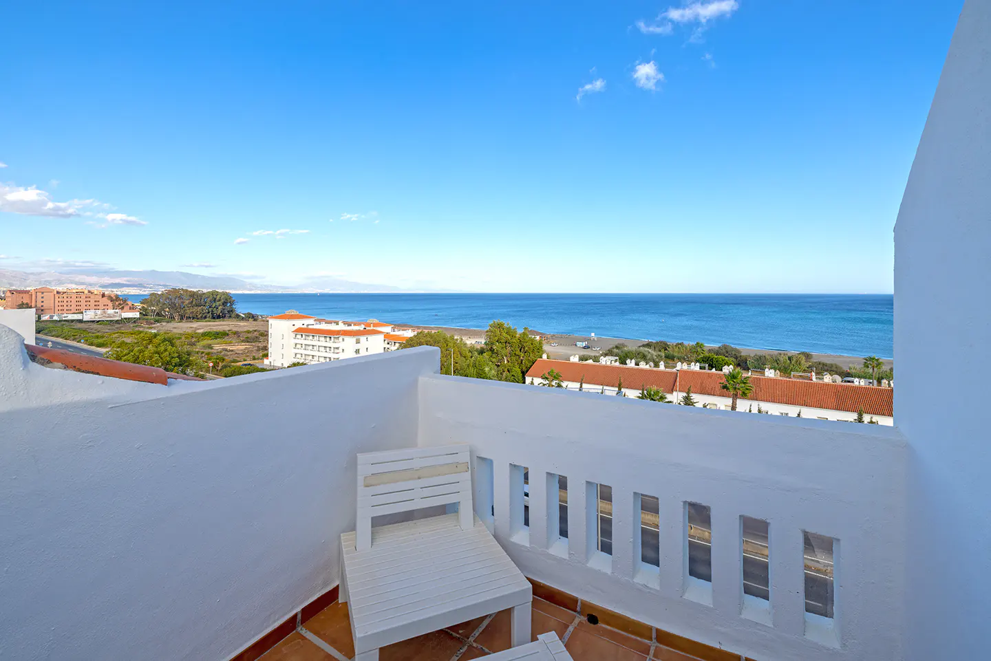 Balcony view with white chair, railing, and tile floor overlooking the ocean and buildings under a blue sky.