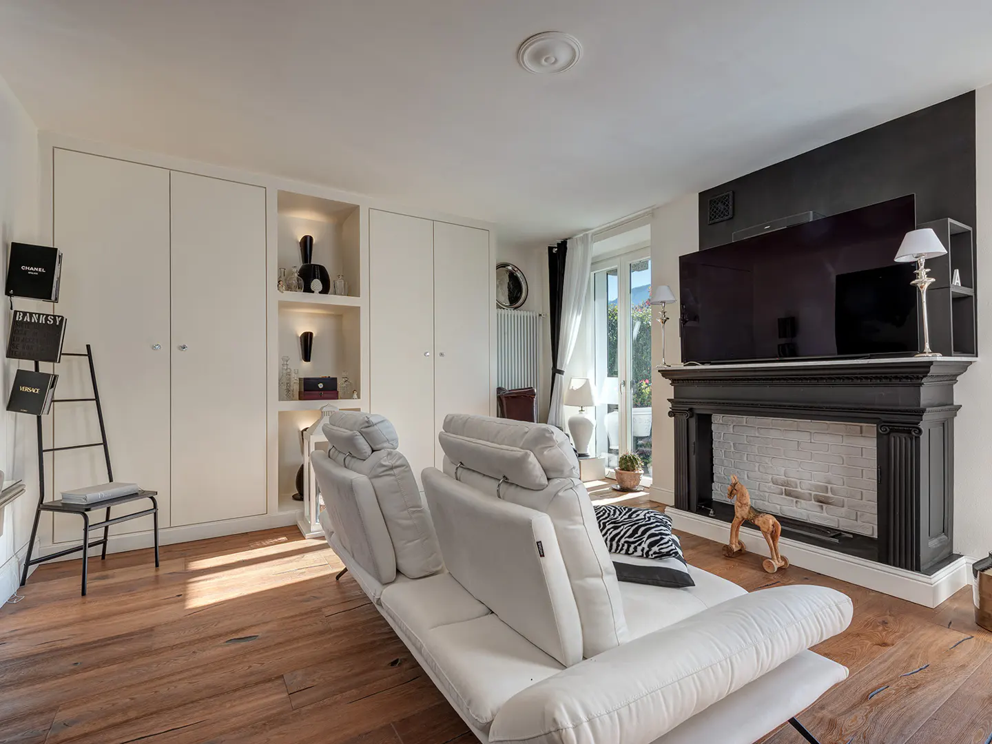 Bright living room with hardwood floors, white walls, and a white leather sofa. A black fireplace with a TV above it is on the right. Built-in white cabinets are on the left.