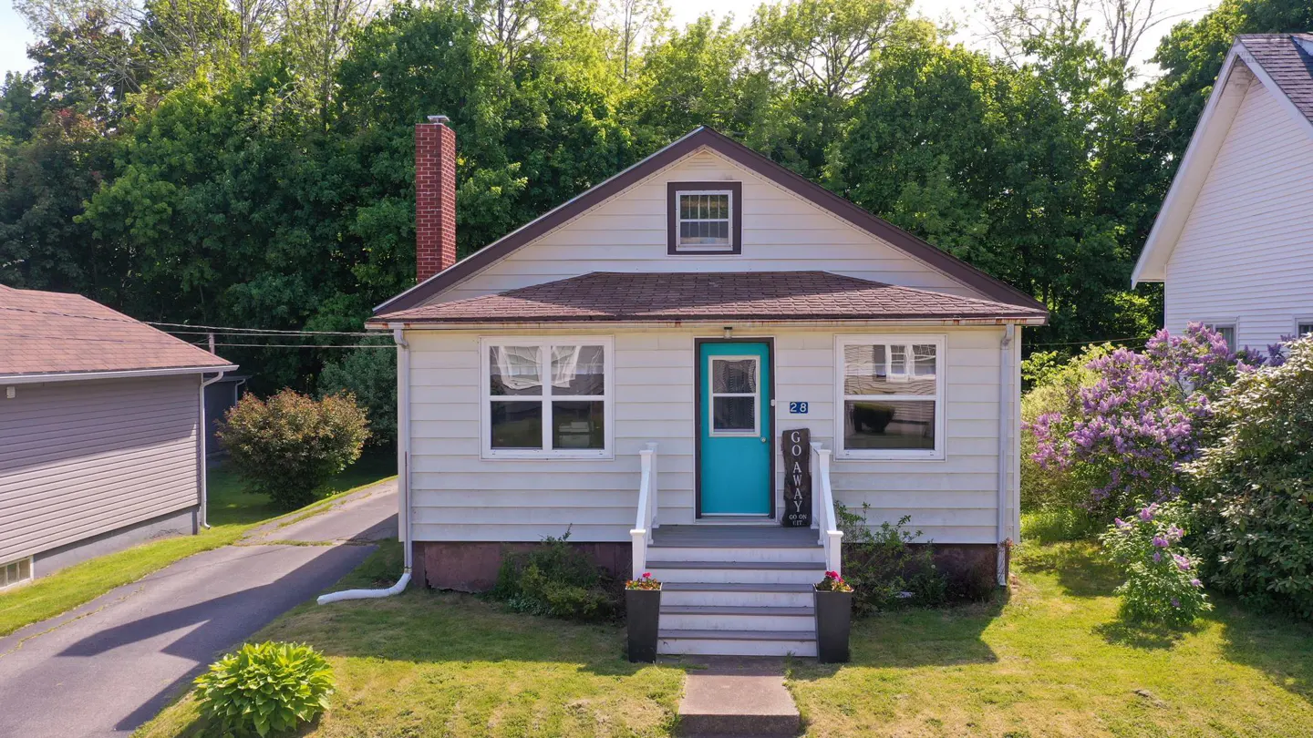 A white single-story house with a bright teal front door, brick chimney, and front steps surrounded by green trees.