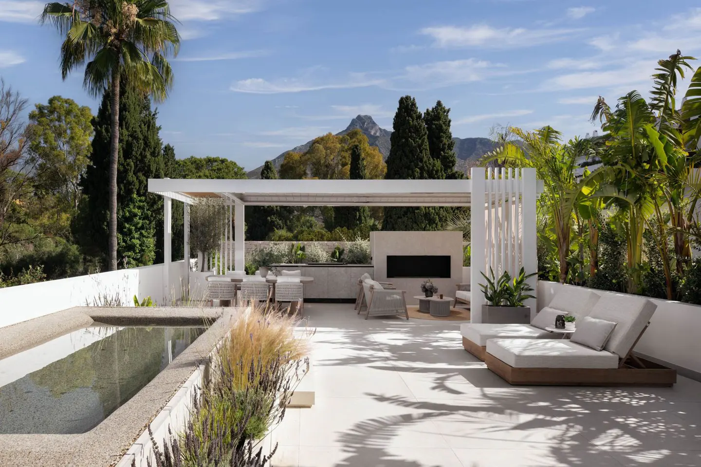 Luxury outdoor living space with pool, pergola, lounge chairs, dining table, and fireplace against a backdrop of lush greenery and mountain views.