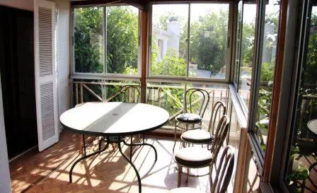 Sunroom with a round white table, black chairs, and large windows overlooking a green garden. White shuttered doors are on the left.