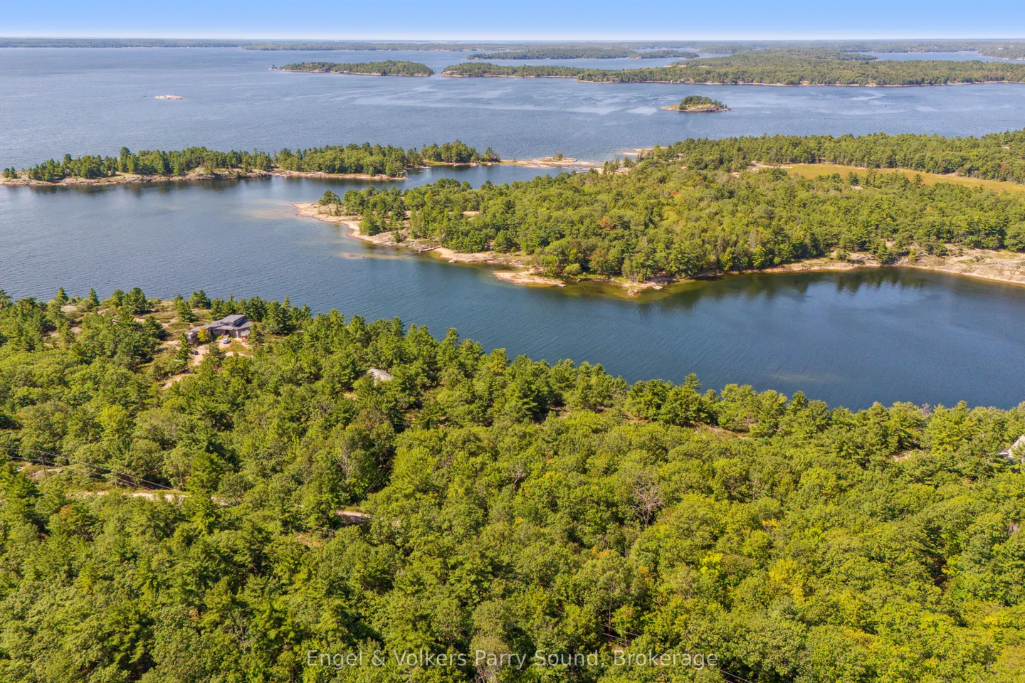 Aerial view of a lake with islands, green trees, and a house on the shore under a blue sky.