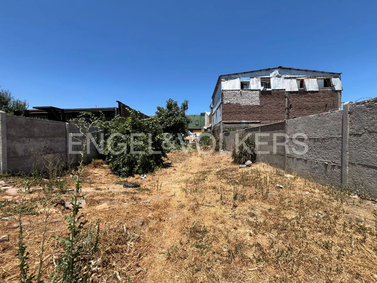 Vacant lot with dry grass, bordered by concrete walls. A building with brick and metal siding is in the background. Blue sky above.