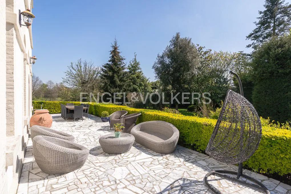 Outdoor patio with wicker furniture, including chairs, a table, and a hanging swing chair, on a stone tile floor with green hedges and trees in the background.