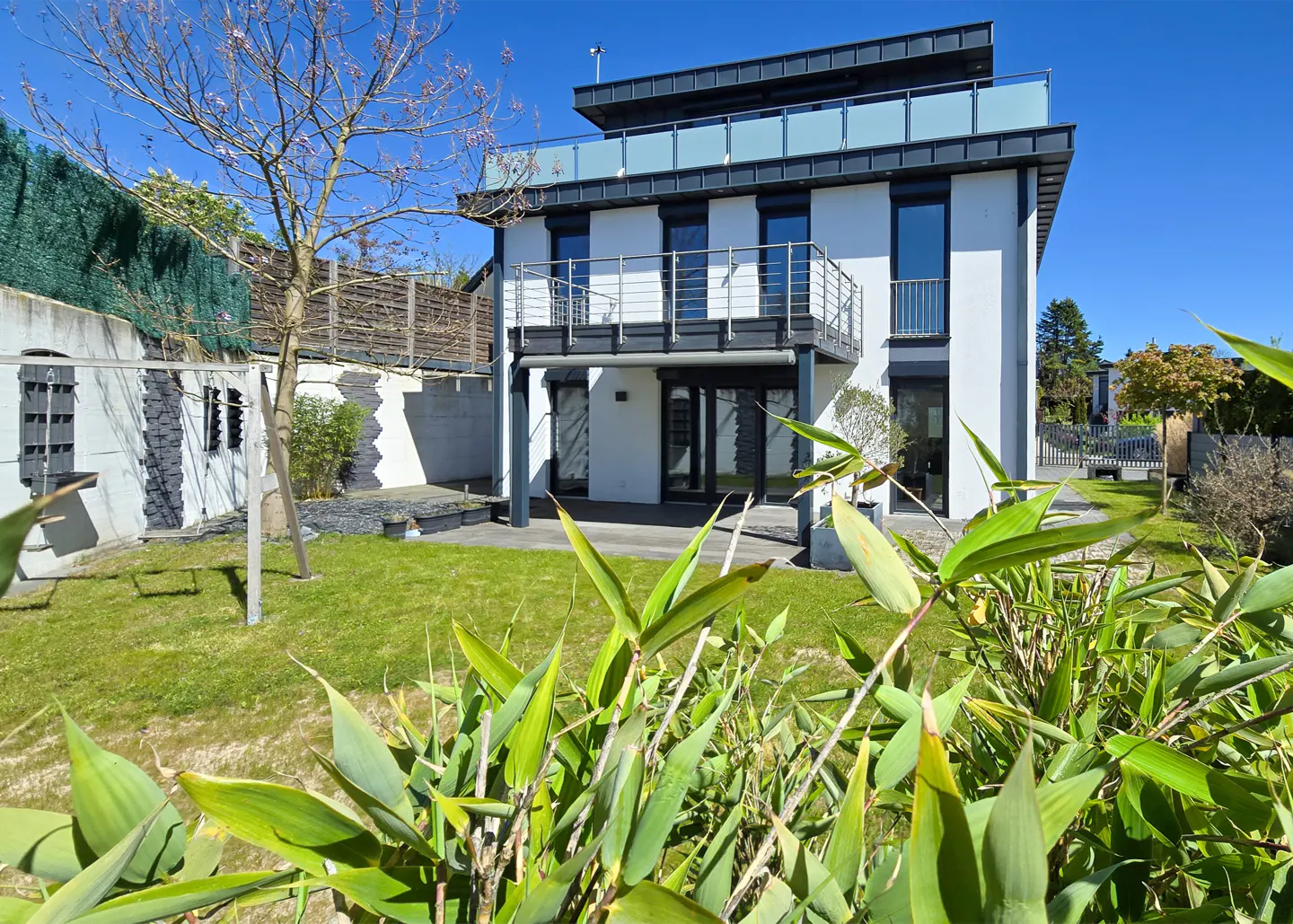 Modern two-story white house with black trim, a balcony, and a green lawn under a clear blue sky.