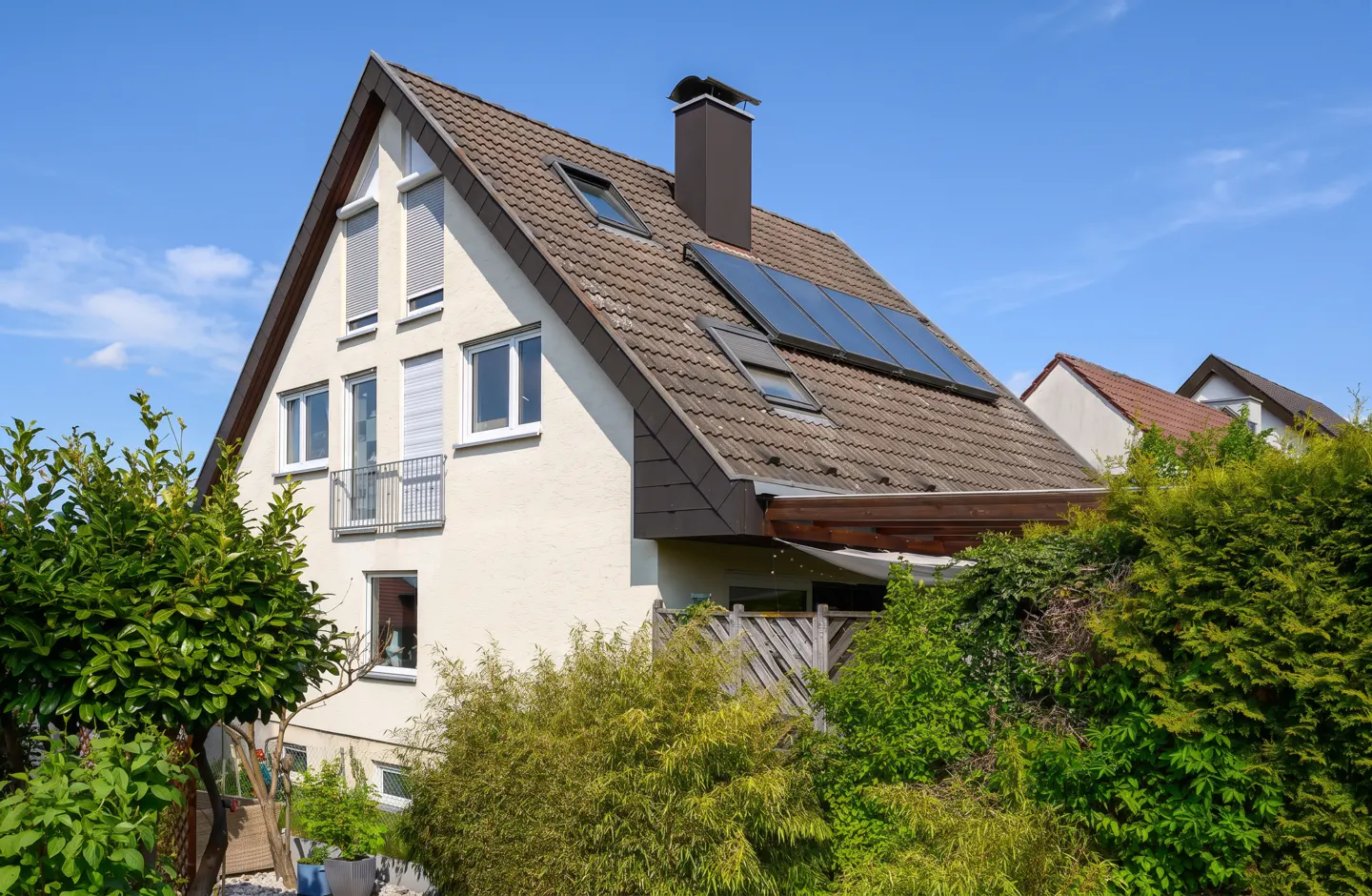 Two-story house with a brown tiled roof, solar panels, and a chimney against a blue sky. Lush green bushes surround the house.