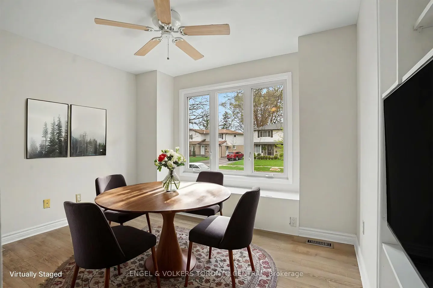 A dining room with a round wooden table, four chairs, and a floral centerpiece. A window overlooks a suburban street.