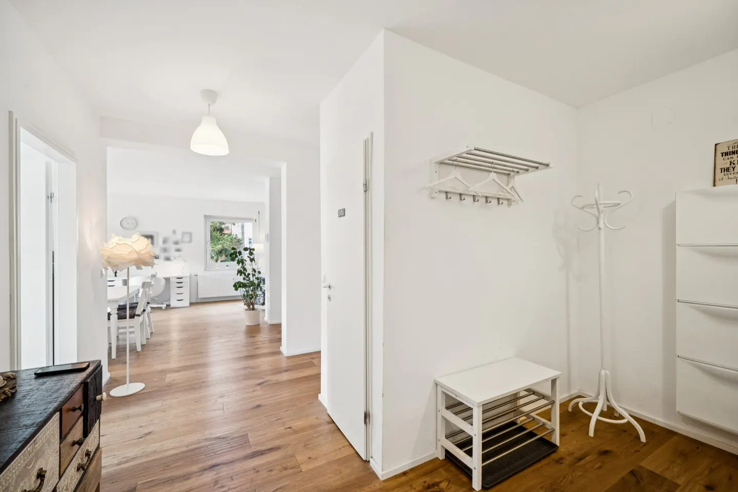 Bright, white hallway with wood floors. A coat rack, shoe bench, and storage cabinet are on the right. A living area is visible through an open doorway.