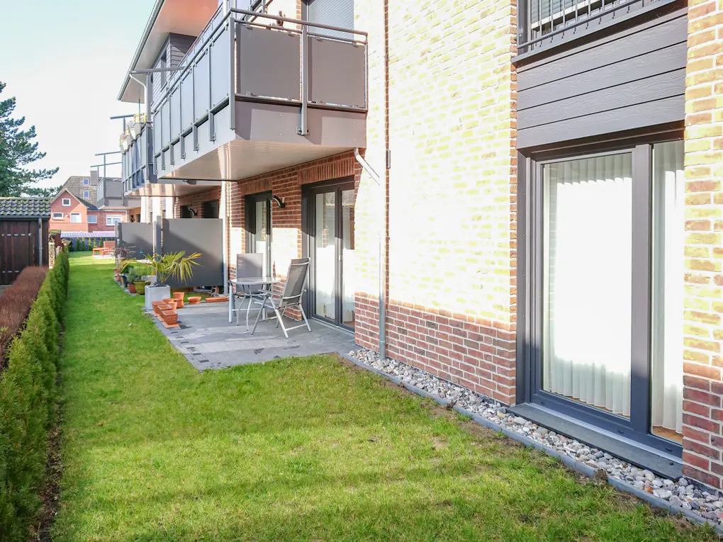 Exterior view of a brick apartment building with balconies, a patio with a table and chairs, and a green lawn.