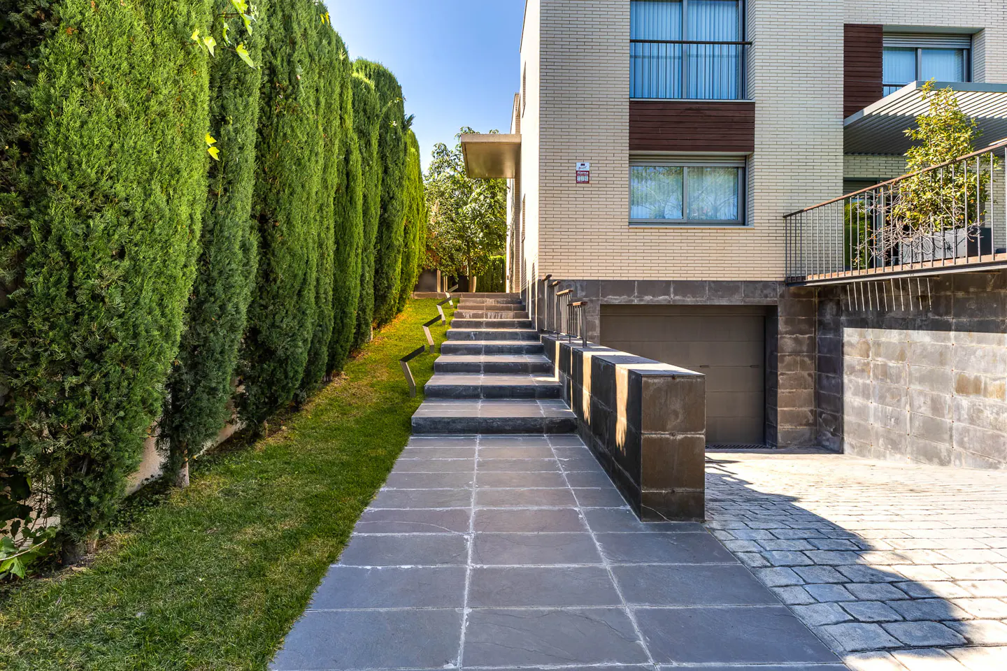 Exterior view of a modern home with a stone path leading to the entrance. Tall green hedges line the left side of the path. A garage is visible on the right.