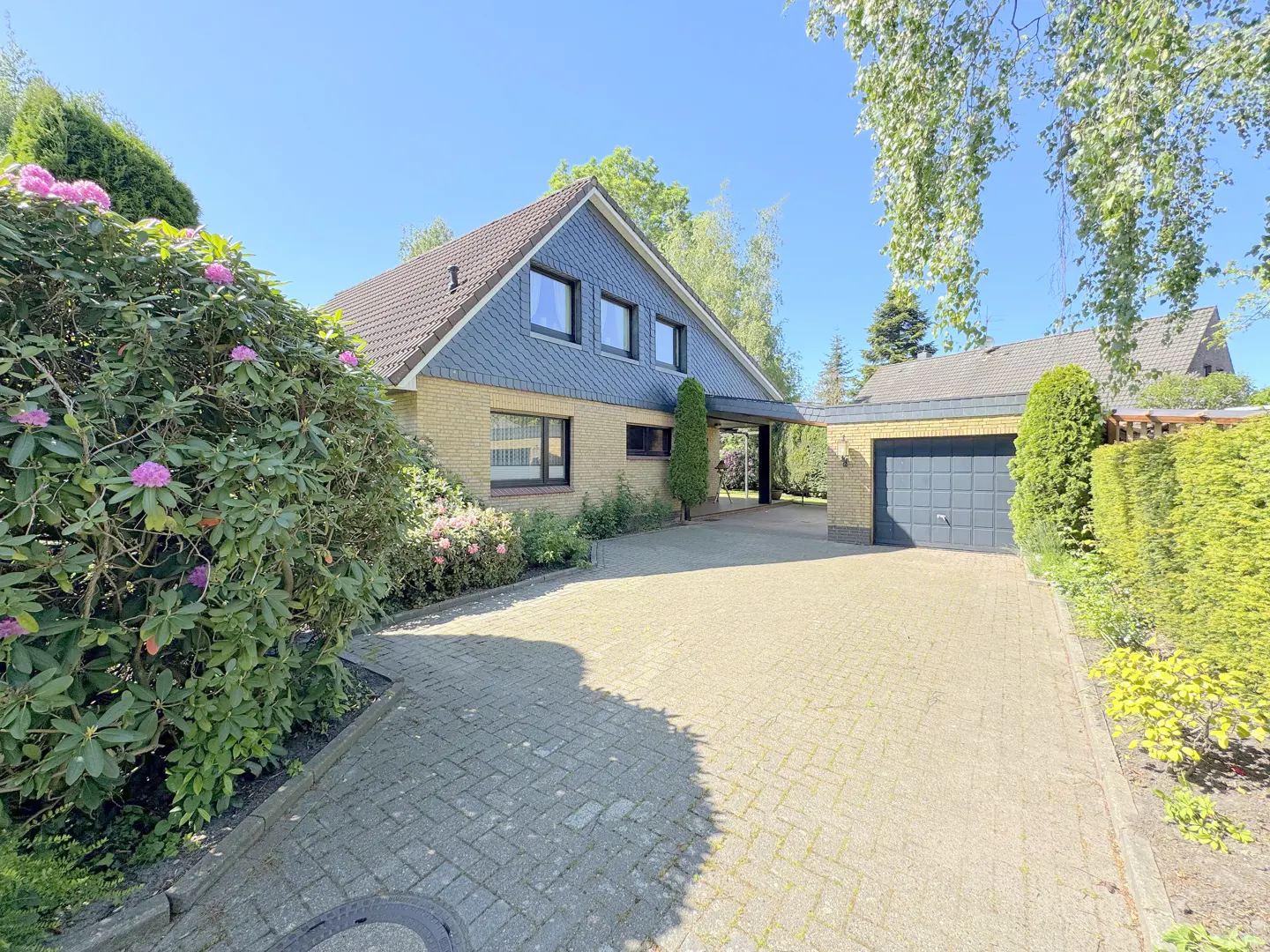 Exterior of a yellow brick house with a gray roof, a driveway, and green bushes.
