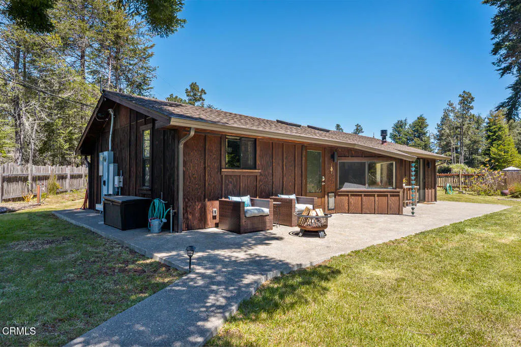 Exterior view of a brown, wood-sided house with a concrete patio, chairs, and a fire pit. Trees and blue sky in the background.