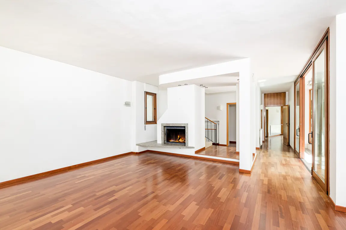 Bright, empty living room with hardwood floors, white walls, and a fireplace with a burning fire. Hallway with doors and stairs visible.