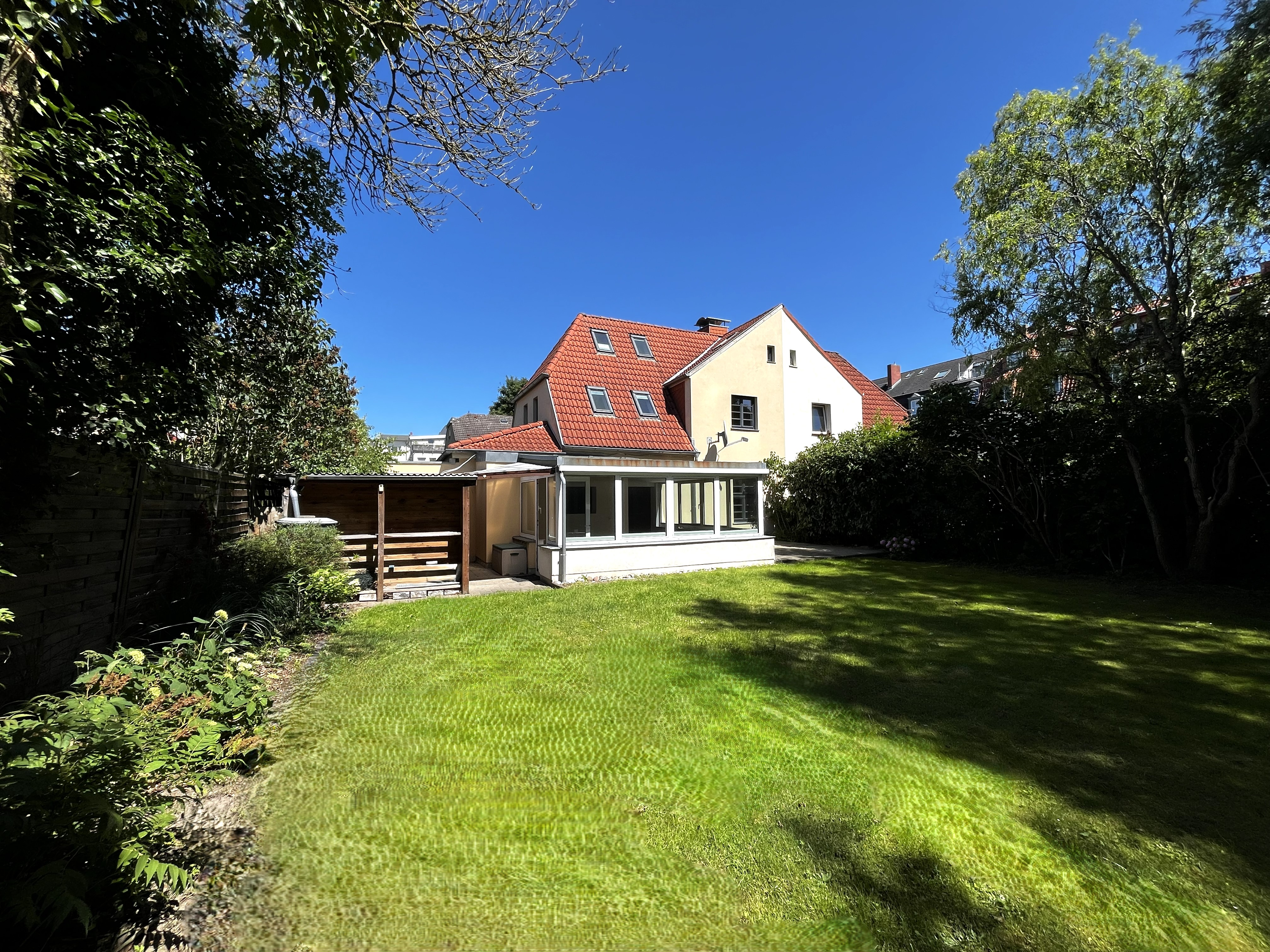 Backyard view of a two-story house with a red tile roof, sunroom, and a large green lawn under a clear blue sky. [AI-generated text]