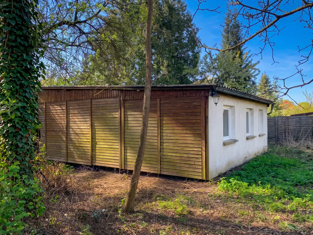 Exterior view of a one-story building with a wooden facade and white walls, surrounded by trees and greenery.