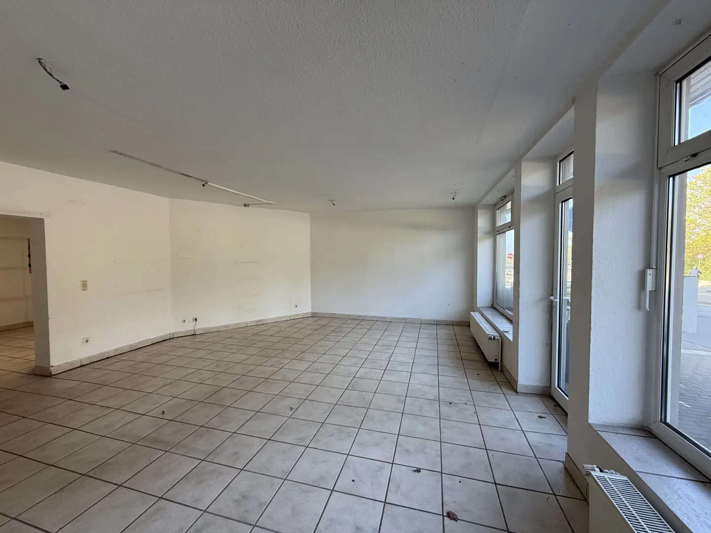 Bright, empty commercial space with white walls, tile floor, and large windows. Radiators sit beneath the windows.