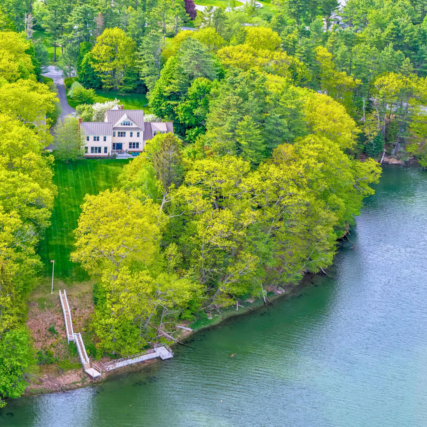 Aerial view of a tan house with a dock on a lake, surrounded by green trees.