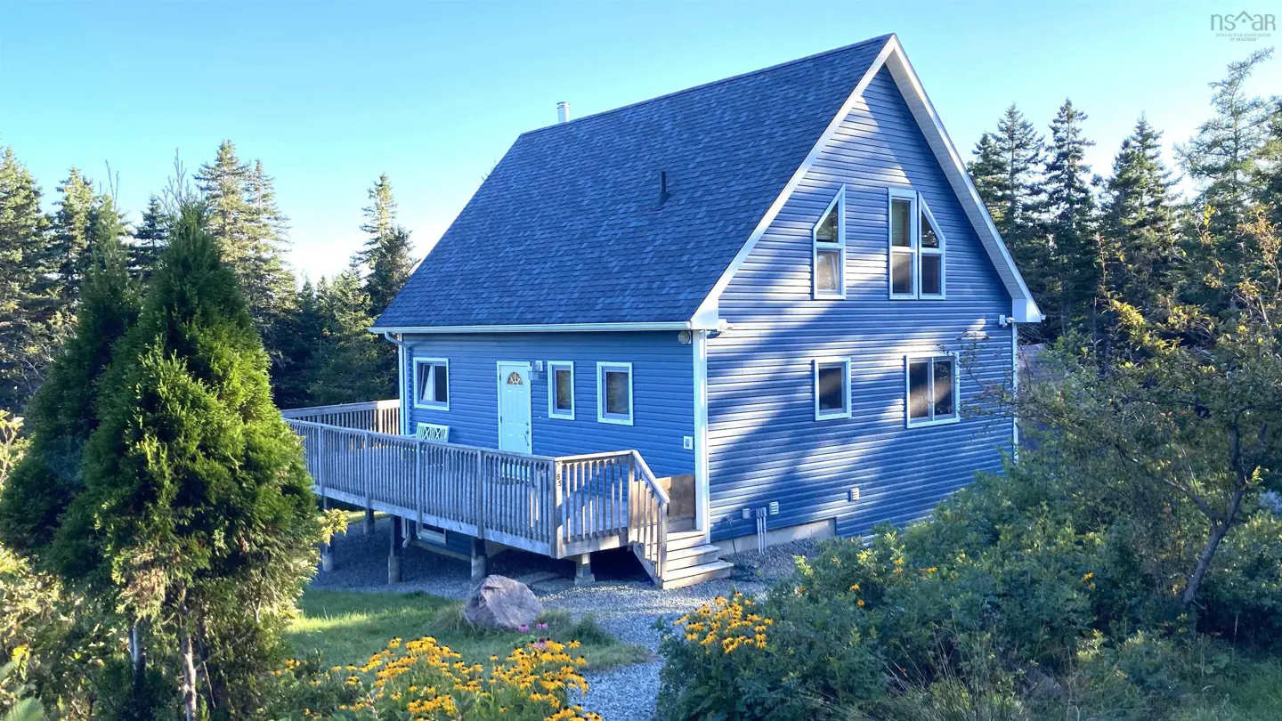 Blue A-frame house with a wooden deck, surrounded by trees and yellow wildflowers.