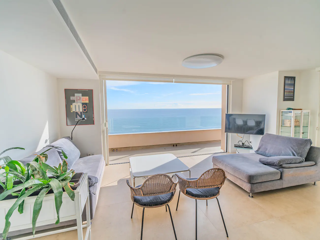 Bright living room with ocean view. Gray sofas, wicker chairs, and a white table. Large sliding doors open to a balcony overlooking the sea.
