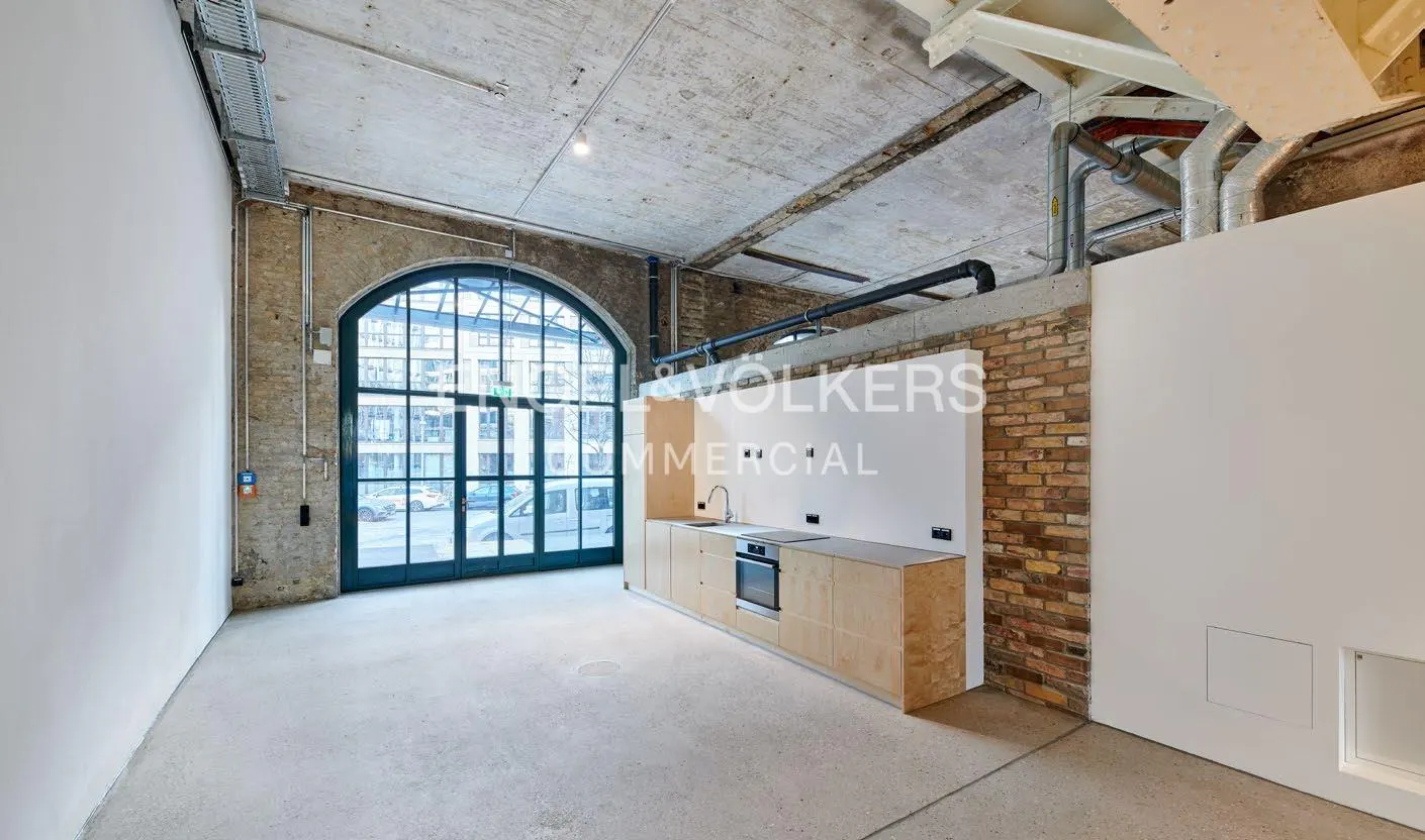 An interior shot of a modern loft apartment with a kitchen, large arched window, and exposed brick and concrete.