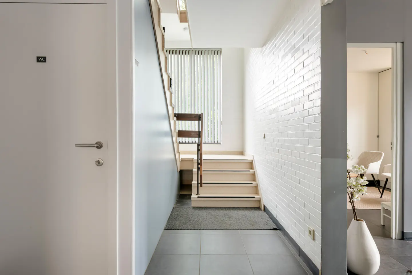 Modern hallway with a white brick wall, light wood staircase, grey tiled floor, and a door labeled WC.