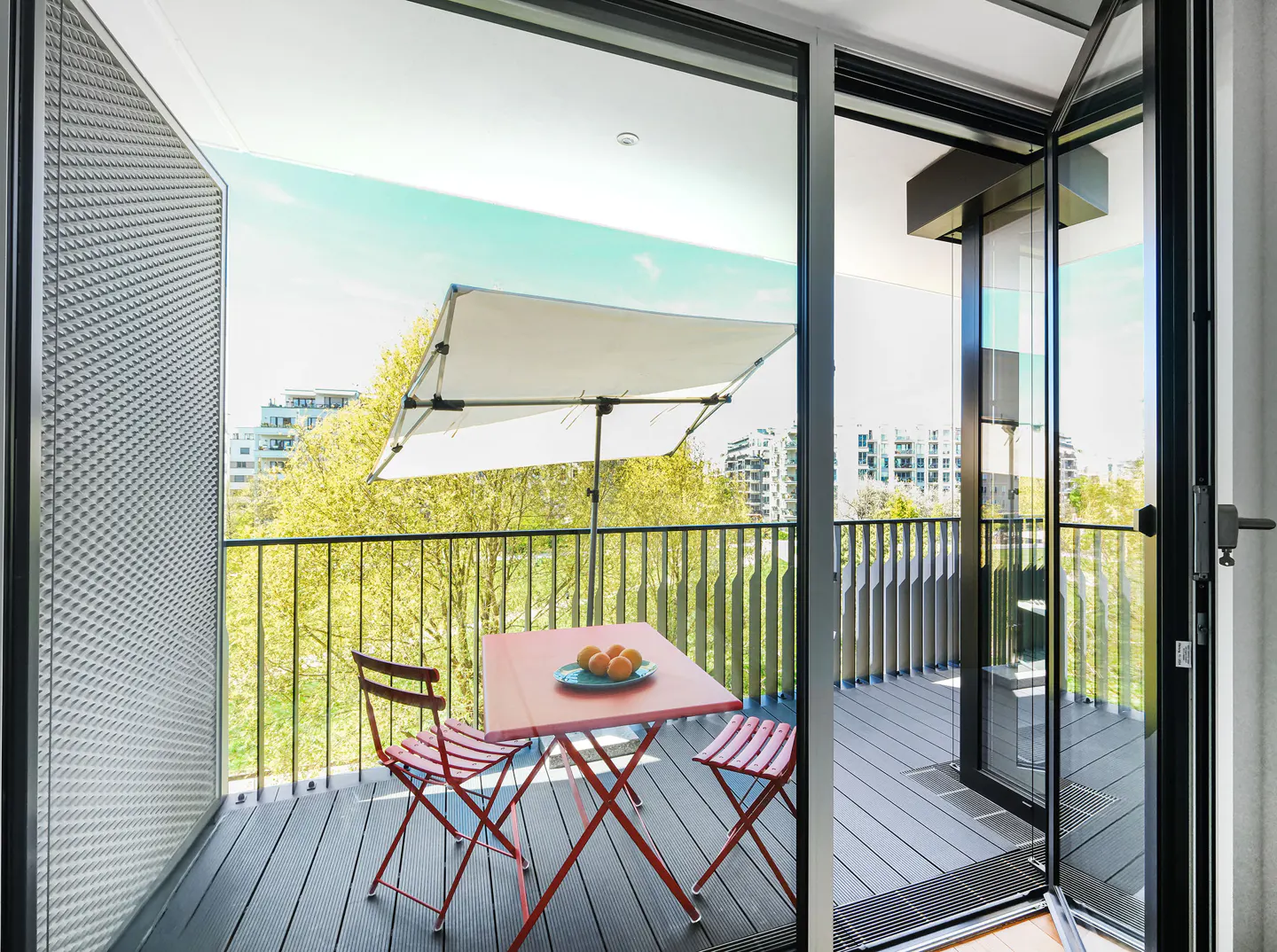 Balcony with red table, chairs, and oranges. White awning, black railing, and gray deck. City view through open glass doors.