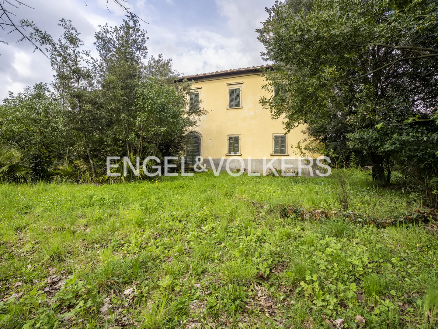 Exterior view of a yellow two-story house with green shutters, surrounded by trees and a grassy lawn. "Engel & Volkers" logo is visible.