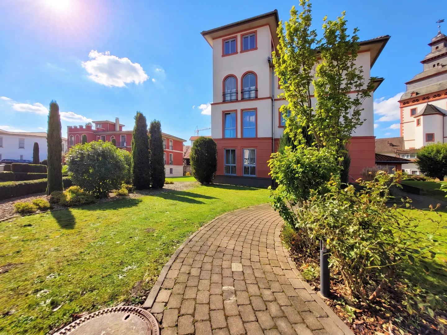 Exterior view of a cream and red building with arched windows, a brick path, and green lawn under a blue sky.