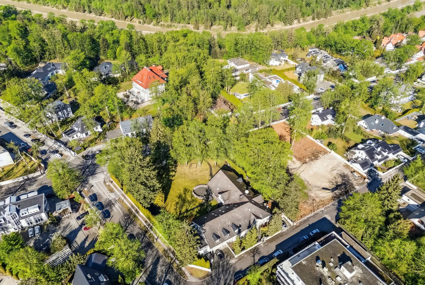 Aerial view of a residential area with houses, trees, and a river in the background. The houses are various sizes and styles, with green trees surrounding them.