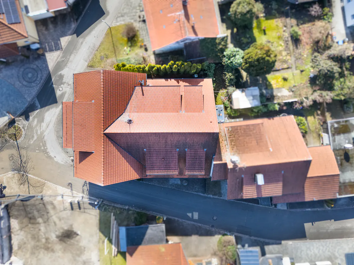 Aerial view of a house with a red tile roof, surrounded by trees and other houses. A road runs alongside the house.