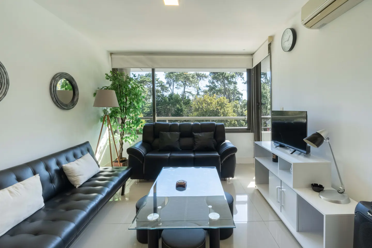 Bright living room with black leather sofas, glass table, and white shelving unit with a TV. Large window shows trees outside.
