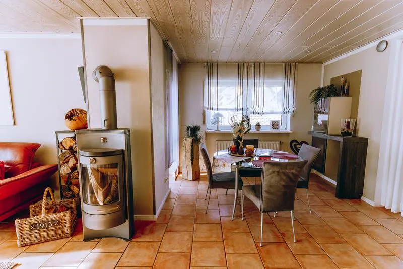 Cozy living room with a wood-burning stove, firewood, and a dining table with chairs on a terracotta tile floor.