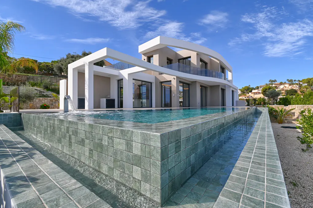 Modern two-story home with a tiled infinity pool under a blue sky. The house is white and gray with large windows and a curved balcony.