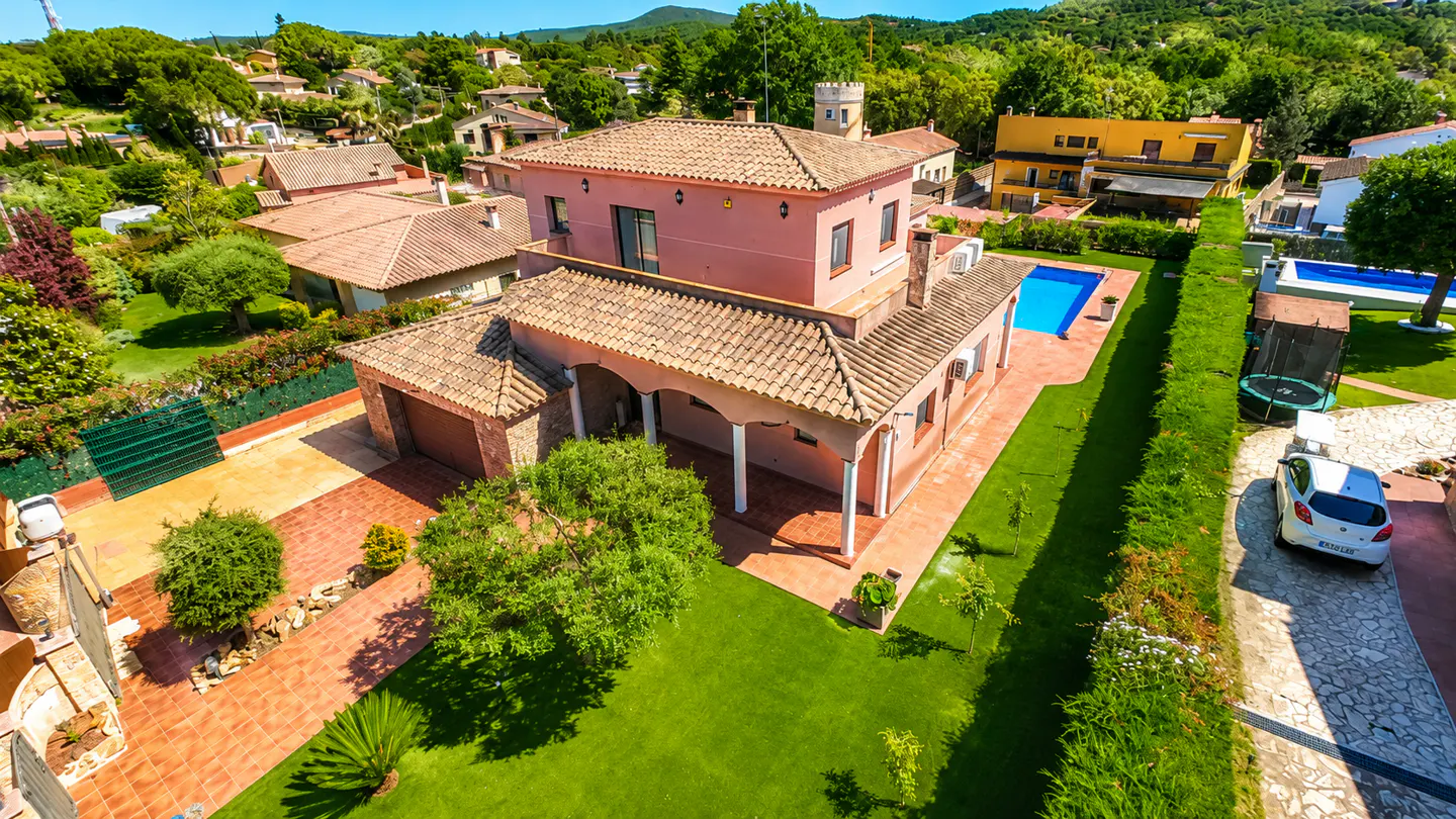 Aerial view of a pink two-story house with a tile roof, green lawn, blue pool, and a white car parked in the driveway.