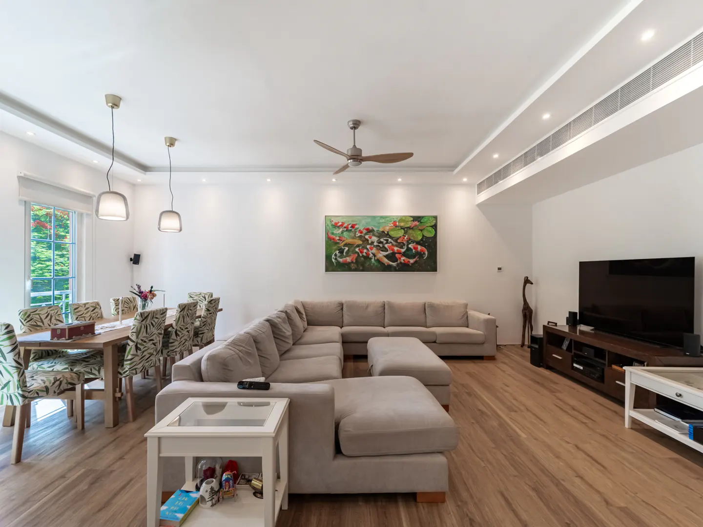 Bright living room with a large beige sectional sofa, wood floors, and a dining table with patterned chairs. A koi fish painting hangs above the sofa.