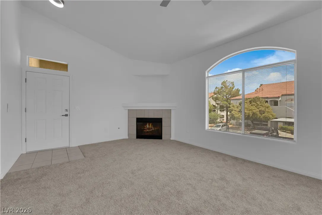 A bright, empty living room with white walls, beige carpet, and a fireplace. A large arched window shows a view of trees and buildings.