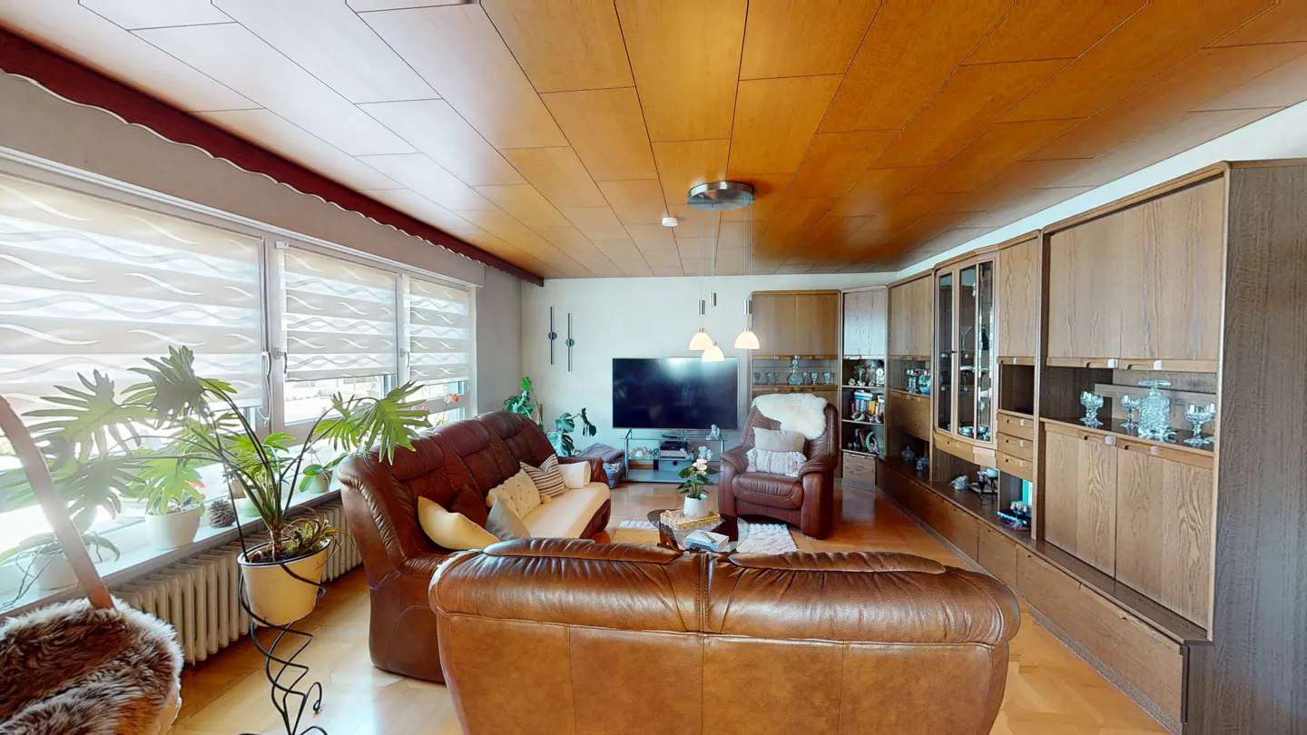 A living room with brown leather sofas, a TV, and built-in wooden cabinets. Large windows provide natural light.