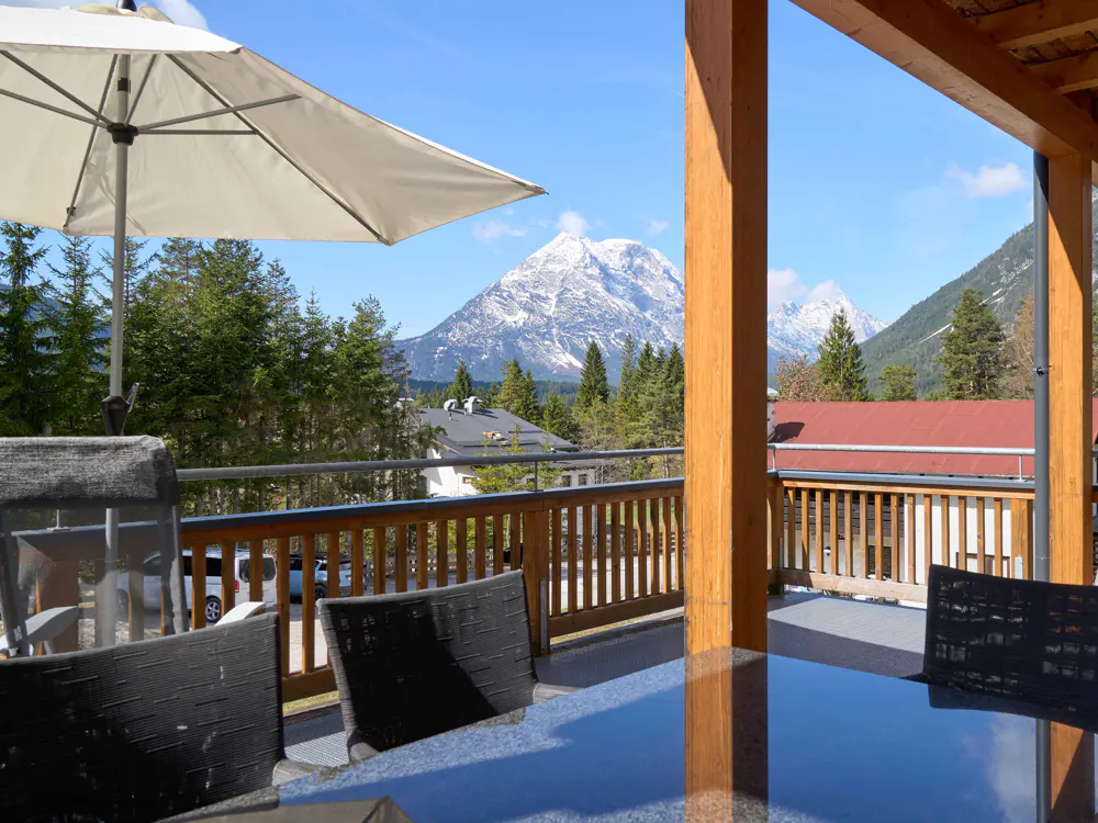 View from a deck with a table, chairs, and umbrella, overlooking trees, houses, and snow-capped mountains under a blue sky.