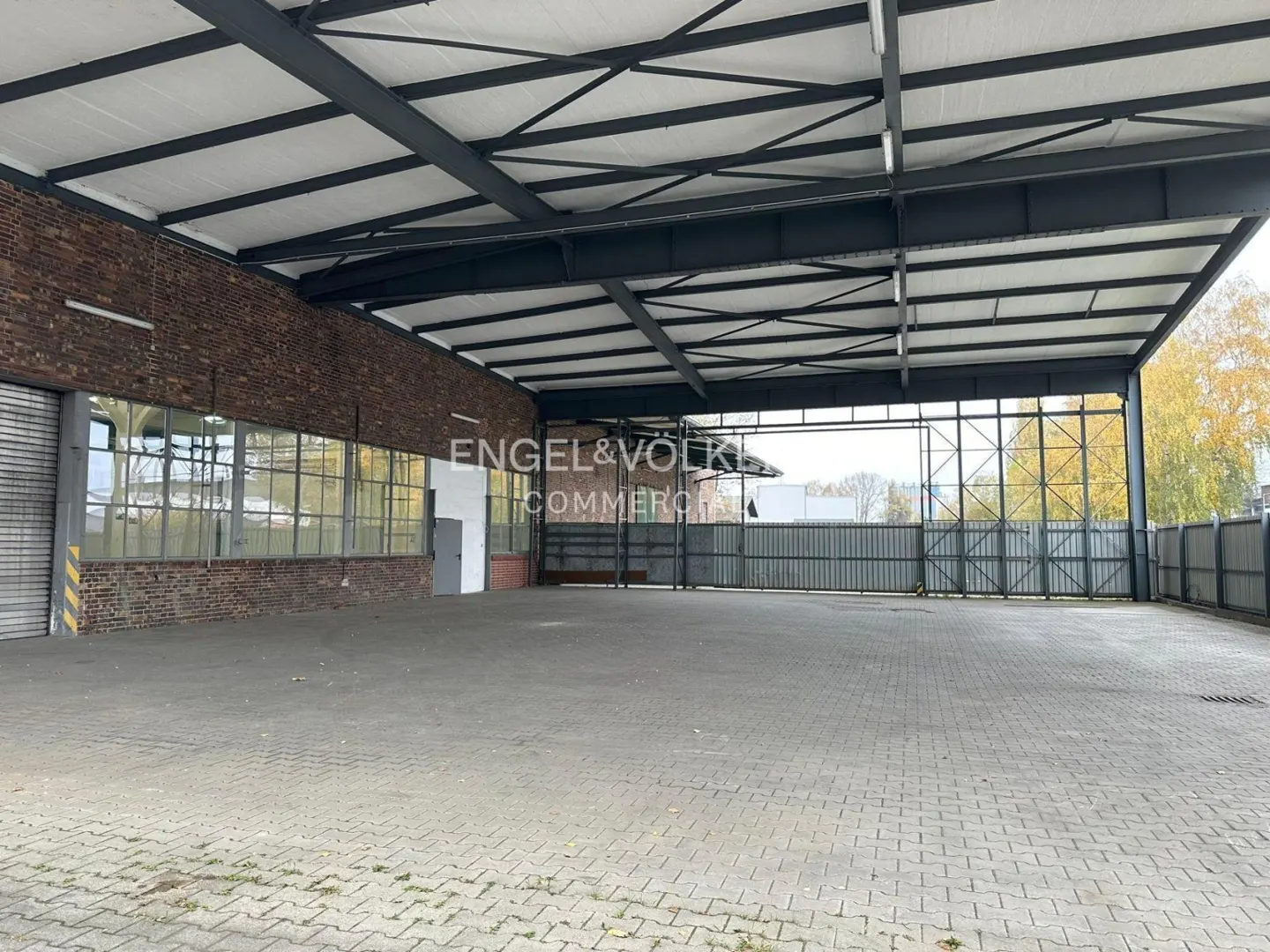 Spacious, open-air industrial warehouse with a paved floor, brick walls, and a high metal roof structure.