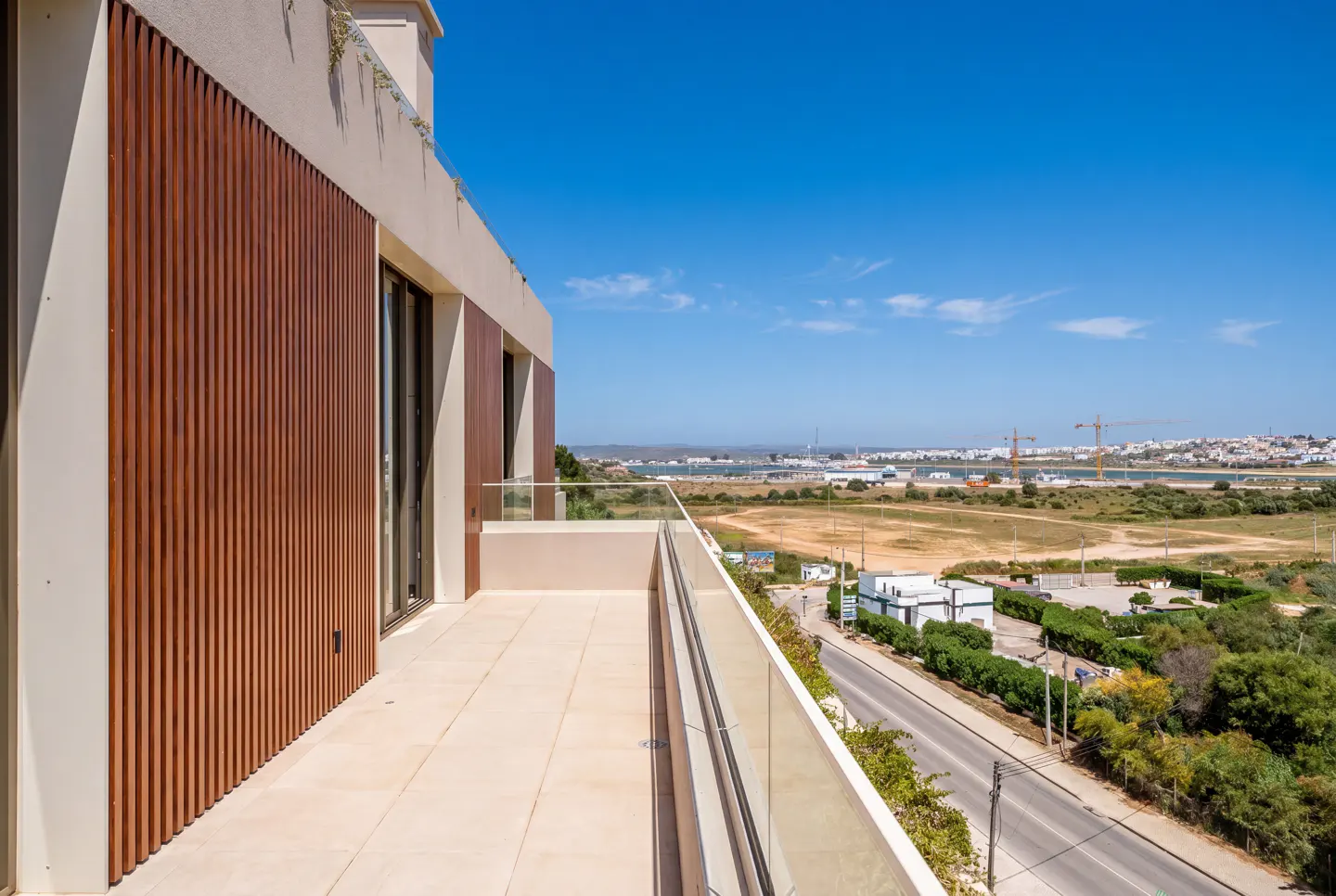 Balcony view of a modern building with wood paneling, glass railing, and a scenic view of the city and harbor under a clear blue sky.