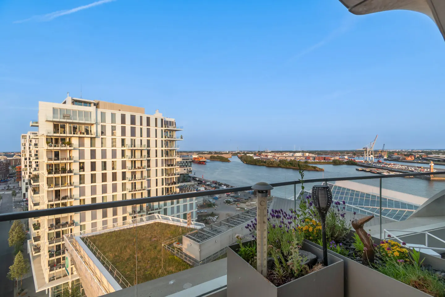 View from a high-rise balcony with potted plants, overlooking a river, city, and modern white building.