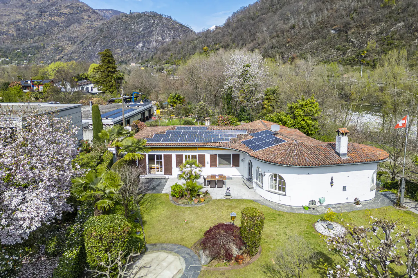 Aerial view of a white house with a red tile roof and solar panels, surrounded by green lawns and trees, with mountains in the background.