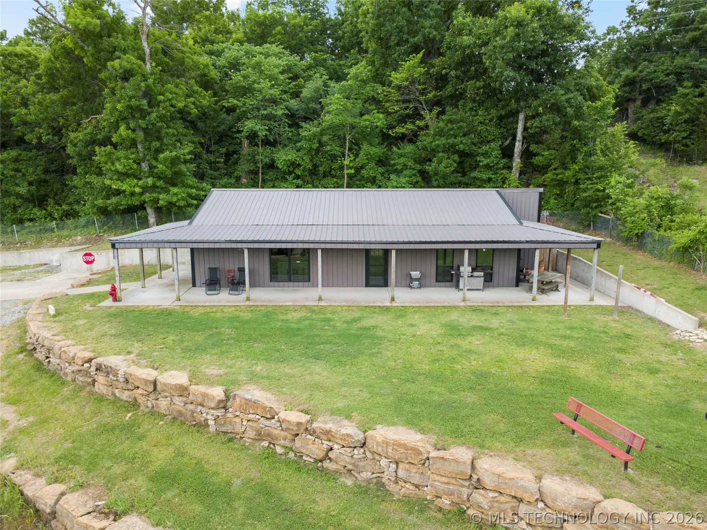 A gray house with a metal roof sits on a green lawn, backed by trees. A stone wall and red bench are in the foreground.