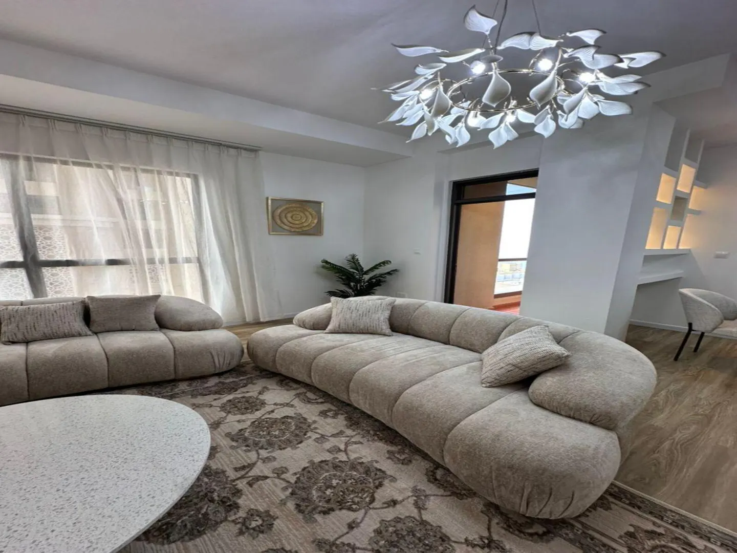 Living room with beige curved sofa, floral rug, and leaf-shaped chandelier. Sheer curtains cover a window, and a balcony is visible in the background.