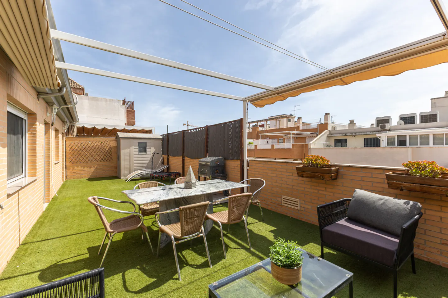 Outdoor patio with green turf, table, chairs, and lounge seating. Brick walls with flower boxes and a shed in the background.